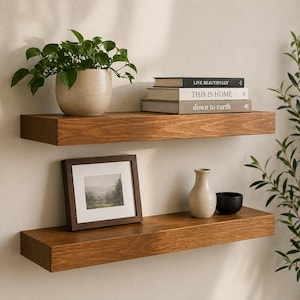 May include: Two floating wooden shelves stained in a warm brown tone. The top shelf holds a potted plant, and books with titles like "LIVE BEAUTIFULLY" and "THIS IS HOME". The bottom shelf displays a framed picture, a vase, and a small black bowl.