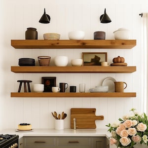 May include: Wooden floating shelves display various kitchen items against a white paneled wall. Items include bowls, plates, mugs, a cutting board, and a small stool. The shelves are illuminated by two black cone-shaped lights.