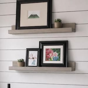 Può includere: Due mensole galleggianti in legno con opere d'arte incorniciate e piccole piante in vaso. La mensola superiore contiene un paesaggio montano incorniciato. La mensola inferiore mostra due immagini incorniciate, una con fiori e l'altra con una persona.
