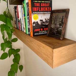 May include: A wooden shelf with a light brown stain, mounted on a white wall. The shelf is holding a stack of books, including "The Great Influenza" by John M. Barry, and a framed black and white photograph.