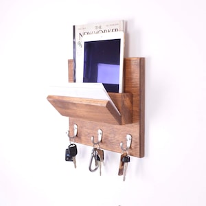 Könnte beinhalten: Ein wandmontierter Schlüsselhalter aus Holz mit einem Regal für Post oder ein Tablet. Der Schlüsselhalter hat vier Haken und ist in einem dunklen Braunton gebeizt. Das Regal ist aus hellerem Holz und hat ein Magazin oder ein Tablet darauf. Der Text auf dem Magazin lautet "The New Yorker".