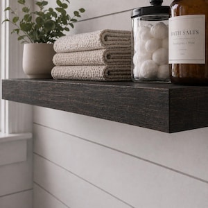 May include: A dark stained wooden shelf displays folded beige towels, a potted plant, a glass jar with cotton balls, and a brown bottle labeled "BATH SALTS Eucalyptus + Mint". The shelf is mounted on a white shiplap wall.