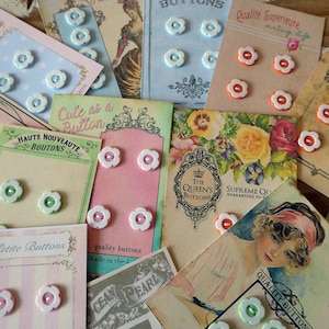 May include: A selection of vintage buttons in a variety of colours and styles, including white, pink, green, and orange. The buttons are displayed on cards with text that reads "Cute as a Button", "Haute Nouveauté Boutons", "The Queen's Buttons", "Supreme Quality Guaranteed to", and "Qualité Supérieure vintage style".