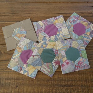 May include: A collection of handmade fabric coasters. Each coaster features a patchwork design with a central hexagon in shades of purple and green. The patchwork includes various pastel colors and patterns, set against a neutral background.