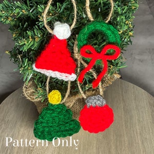Puede incluir: Cuatro adornos navideños de crochet colgando de un pequeño árbol de Navidad artificial. Los adornos son un gorro de Papá Noel rojo y blanco, una corona verde con un lazo rojo, un árbol de Navidad verde y un adorno rojo con una parte superior gris.