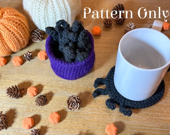 Può includere: Un calderone a uncinetto viola riempito di filato nero, un sottobicchiere a uncinetto nero a forma di ragno con una tazza bianca sopra e una superficie di legno con oggetti decorativi arancioni e marroni. Il testo "Pattern Only" è visibile nell'immagine.