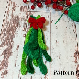 May include: A crocheted green and red mistletoe sprig with a red bow. The mistletoe is on a white wooden background with a strand of red berries and green balls of yarn.