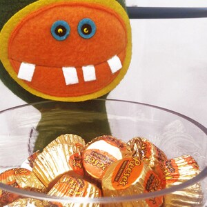 May include: A green felt monster with an orange felt face and white teeth holds a glass bowl filled with Reese's Peanut Butter Cups wrapped in gold foil.