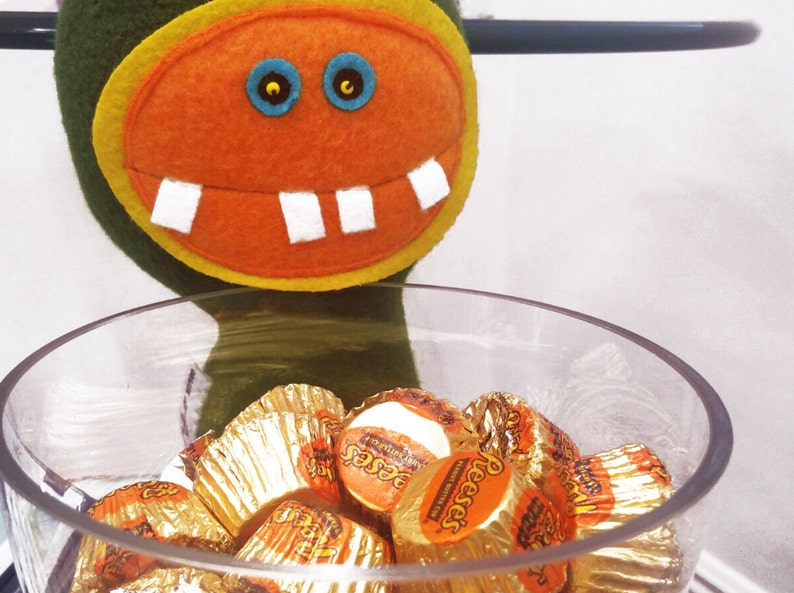 May include: A green felt monster with an orange felt face and white teeth holds a glass bowl filled with Reese's Peanut Butter Cups wrapped in gold foil.