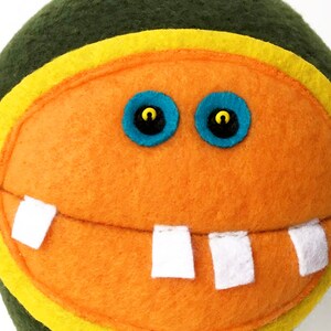 May include: A plush toy with a green, yellow, and orange face. The toy has two blue eyes with black pupils and a wide, toothy smile.