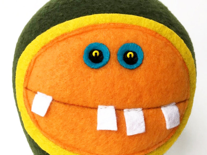 May include: A plush toy with a green, yellow, and orange face. The toy has two blue eyes with black pupils and a wide, toothy smile.