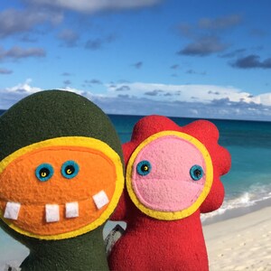May include: Two plush toys with smiling faces stand on a white sandy beach with a blue ocean in the background. The toy on the left is green with a yellow and orange face. The toy on the right is red with a pink face.