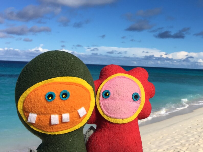 May include: Two plush toys with smiling faces stand on a white sandy beach with a blue ocean in the background. The toy on the left is green with a yellow and orange face. The toy on the right is red with a pink face.
