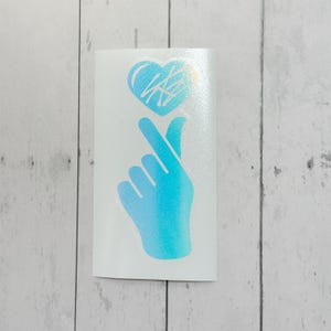 May include: A white sticker with a hand gesture forming a heart shape. The hand and heart are light blue with a holographic effect. The heart has scribbled lines inside.