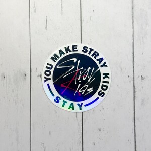 May include: A circular, holographic sticker featuring the text "YOU MAKE STRAY KIDS STAY". The words "Stray Kids" are in a stylized font, with blue and red accents. The sticker has a reflective, iridescent finish, creating a rainbow effect.