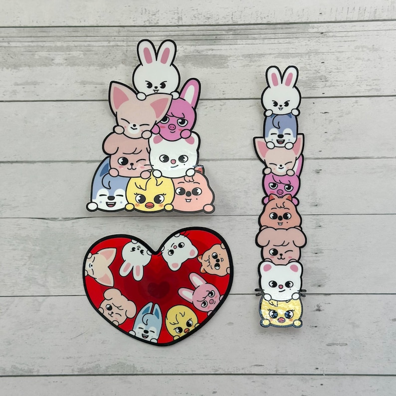 May include: A collection of colourful stickers featuring cartoon animal characters. The stickers include a stack of animals, a tall tower of animals, and a heart-shaped design filled with various animal faces. The stickers are on a light grey wooden surface.