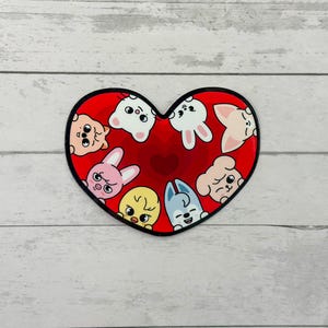 May include: A heart-shaped sticker with a red background and a black border. The sticker features cartoon animal faces in various colours, including white, pink, yellow, and blue. The animals are arranged around the heart's centre.