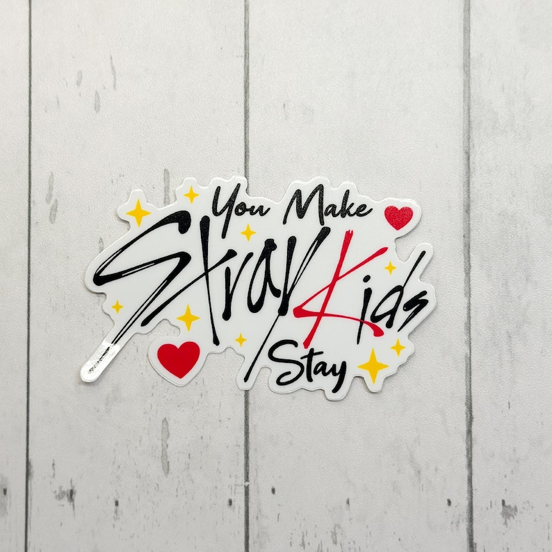 May include: A white sticker featuring the text "You Make Stray Kids Stay" in black and red script. The sticker has a whimsical design with yellow stars and red hearts. The sticker is on a white and grey surface.