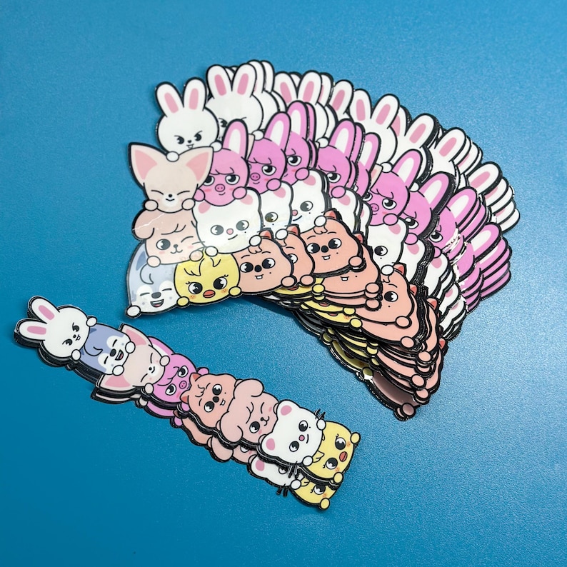 May include: A collection of colourful stickers featuring cartoon animal characters, including bunnies, foxes, and bears. The stickers are in various shades of pink, white, and yellow, with black outlines. The stickers are stacked on a blue surface.