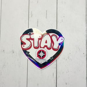 May include: Heart-shaped sticker with the word "STAY" in red, outlined in white and black. The sticker features a compass design and star accents. The sticker has a holographic, iridescent border.