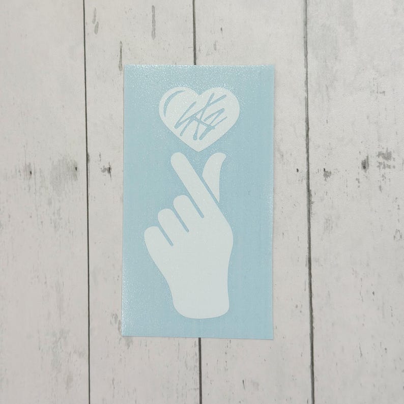 May include: A white vinyl sticker showing a hand gesture forming a heart shape with a heart symbol above the fingers. The sticker has a light blue background and is displayed on a wooden surface.