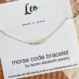 May include: A delicate silver Morse code bracelet with pale green beads spelling "Leo". The bracelet is displayed on a card with the word "Leo" and the Morse code dots and dashes. The card also includes the text "morse code bracelet".