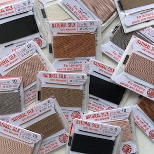 May include: Assortment of natural silk thread spools in various colors, including black, brown, and beige. Each spool is labeled "NATURAL SILK" with the length, number, and diameter of the thread. The spools are designed for stringing pearls and beads.