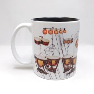 May include: White ceramic mug with a black rim and a design of a drum set with cymbals, timpani, and a snare drum. The design is in black and white with orange and brown accents.