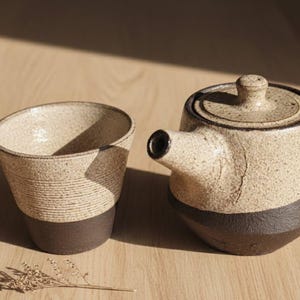 Può includere: Un set da tè in ceramica nei toni del beige e del marrone. Il set comprende una teiera con coperchio e beccuccio e una tazza abbinata. La tazza presenta un design strutturato e a righe. La teiera e la tazza sono su una superficie di legno.