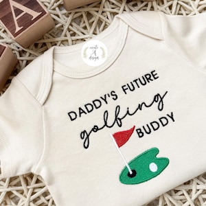 May include: Cream-colored baby onesie with the text "DADDY'S FUTURE golfing BUDDY" in black script. A green golf course design with a red flag is embroidered on the front. Wooden blocks spell "BABY".