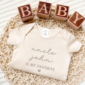 May include: A beige baby onesie with the text "uncle john IS MY FAVORITE" in gray script. The onesie is on a woven basket with wooden blocks spelling "BABY". A small sticker with the text "create A design" is on the onesie.