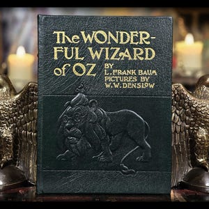 Puede incluir: Un ejemplar antiguo de "The Wonderful Wizard of Oz" de L. Frank Baum, con letras doradas y una cubierta verde oscuro con una ilustraci&oacute;n de un le&oacute;n. A ambos lados del libro, dos sujetalibros de &aacute;guila de bronce crean una exhibici&oacute;n literaria cl&aacute;sica.