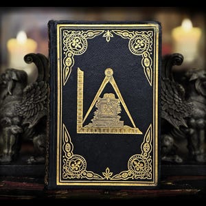 Könnte beinhalten: Ein dunkelblaues Buch mit goldgeprägten Freimaurersymbolen, darunter Winkel, Zirkel und Waage. Der Einband hat verzierte goldene Ränder und ist vor einem verschwommenen Hintergrund mit Kerzen und Statuen platziert.