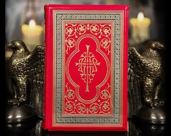 May include: A hardcover book with a vibrant red cover featuring intricate gold foil stamping. The design includes a central ornate emblem surrounded by symmetrical floral scrollwork and a geometric border pattern in black and gold.
