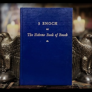 May include: A blue hardcover book with the title "3 Enoch or The Hebrew Book of Enoch" in silver lettering. Two bronze eagle bookends frame the book, with candles softly lit in the background.
