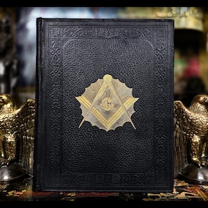 Könnte beinhalten: Ein schwarzes, geprägtes Buch mit einem goldenen Emblem, das ein Quadrat- und Zirkel-Design zeigt. Das Buch wird von zwei goldenen Adlerstatuen flankiert. Der Einband hat einen dekorativen Rand und ein strukturiertes Aussehen, was auf einen antiken oder Vintage-Artikel hindeutet.