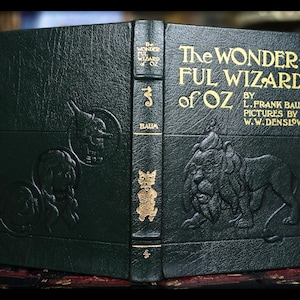 Puede incluir: Un libro verde oscuro en relieve titulado "The Wonderful Wizard of Oz" de L. Frank Baum. La portada presenta letras doradas e ilustraciones en relieve de personajes, incluyendo el Hombre de Hojalata, el Le&oacute;n y el Espantap&aacute;jaros. Una novela de fantas&iacute;a cl&aacute;sica.