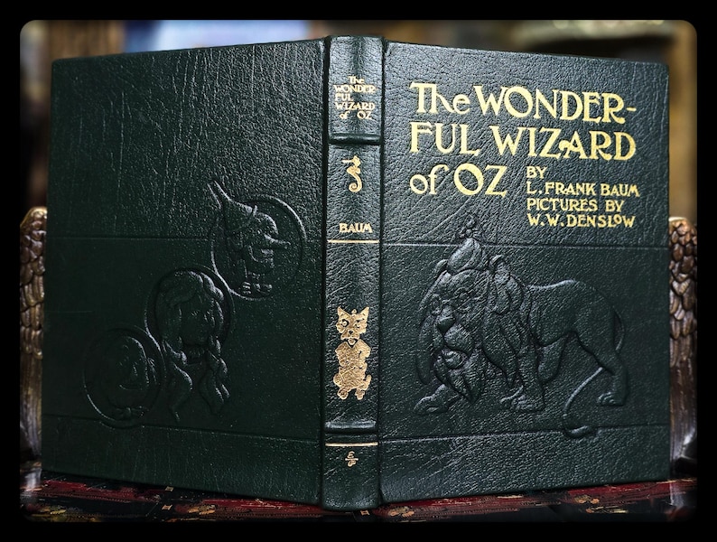 Puede incluir: Un libro verde oscuro en relieve titulado "The Wonderful Wizard of Oz" de L. Frank Baum. La portada presenta letras doradas e ilustraciones en relieve de personajes, incluyendo el Hombre de Hojalata, el Le&oacute;n y el Espantap&aacute;jaros. Una novela de fantas&iacute;a cl&aacute;sica.