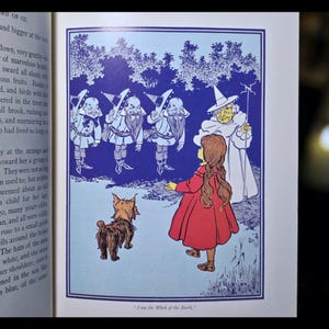Puede incluir: Ilustraci&oacute;n de un libro infantil con una ni&ntilde;a con un vestido rojo, un perro y varias figuras con trajes azules y blancos. El texto en la parte inferior dice "I am the Witch of the North."