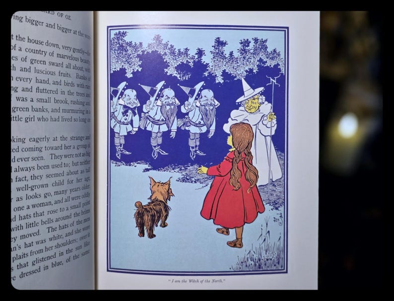 Puede incluir: Ilustraci&oacute;n de un libro infantil con una ni&ntilde;a con un vestido rojo, un perro y varias figuras con trajes azules y blancos. El texto en la parte inferior dice "I am the Witch of the North."