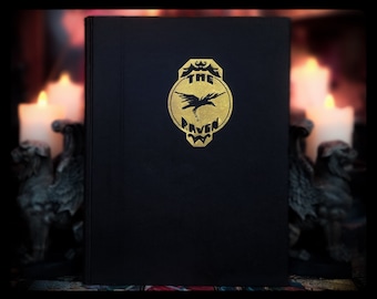 Può includere: Un libro nero con scritte dorate e un emblema dorato raffigurante un corvo e le parole "The Raven". Il libro è in piedi, con candele accese e una statua decorativa sullo sfondo.