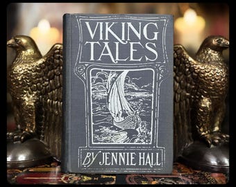 May include: A vintage book titled "VIKING TALES" by Jennie Hall, with a detailed illustration of a Viking ship on the cover. Flanking the book are two bronze eagle statues, creating a historical and decorative display.