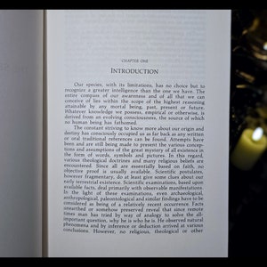 Pode incluir: Uma p&aacute;gina de um livro intitulado "Introdu&ccedil;&atilde;o" com texto sobre a origem e o destino da humanidade. O texto discute as limita&ccedil;&otilde;es do conhecimento humano e a busca por respostas a quest&otilde;es fundamentais sobre a exist&ecirc;ncia. A p&aacute;gina &eacute; iluminada por uma vela.
