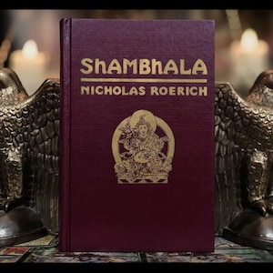 May include: A burgundy hardcover book with gold lettering titled "Shambhala" by Nicholas Roerich. The book is sitting on a table with two golden eagle bookends.