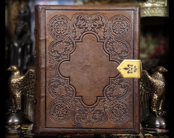 May include: A brown leather-bound book with ornate embossed floral designs and a gold-colored clasp. The cover features a central rectangular panel and decorative corner elements. The book is a vintage style, with a classic aesthetic.