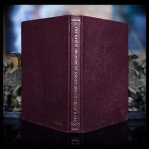 May include: A deep burgundy hardcover book with a textured finish. The spine features gold lettering that reads MALAK, THE MYSTIC GRIMOIRE OF MIGHTY SPELLS AND RITUALS, and PARKER, accompanied by simple gold line graphics at the top and bottom.