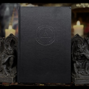 May include: A black book with embossed circular design and a triangle symbol. The book is flanked by two gargoyle statues. The book's cover has a textured appearance. The overall theme is gothic and occult.