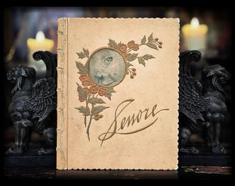 Può includere: Copertina di libro vintage dai toni seppia, con un ritratto ovale di donna e decorazioni floreali arancioni e verdi. La parola "Senore" è scritta in elegante corsivo. Due statue di grifoni neri affiancano il libro.