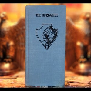 May include: A blue book with the title "The Herbalist" printed on the cover. The cover features a black and white illustration of a Native American wearing a headdress inside a shield.
