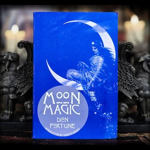 May include: A blue book titled "Moon Magic" by Dion Fortune. The cover features a white crescent moon with a seated figure and the book's title in a white circle. The book is displayed between two dark gray statues.